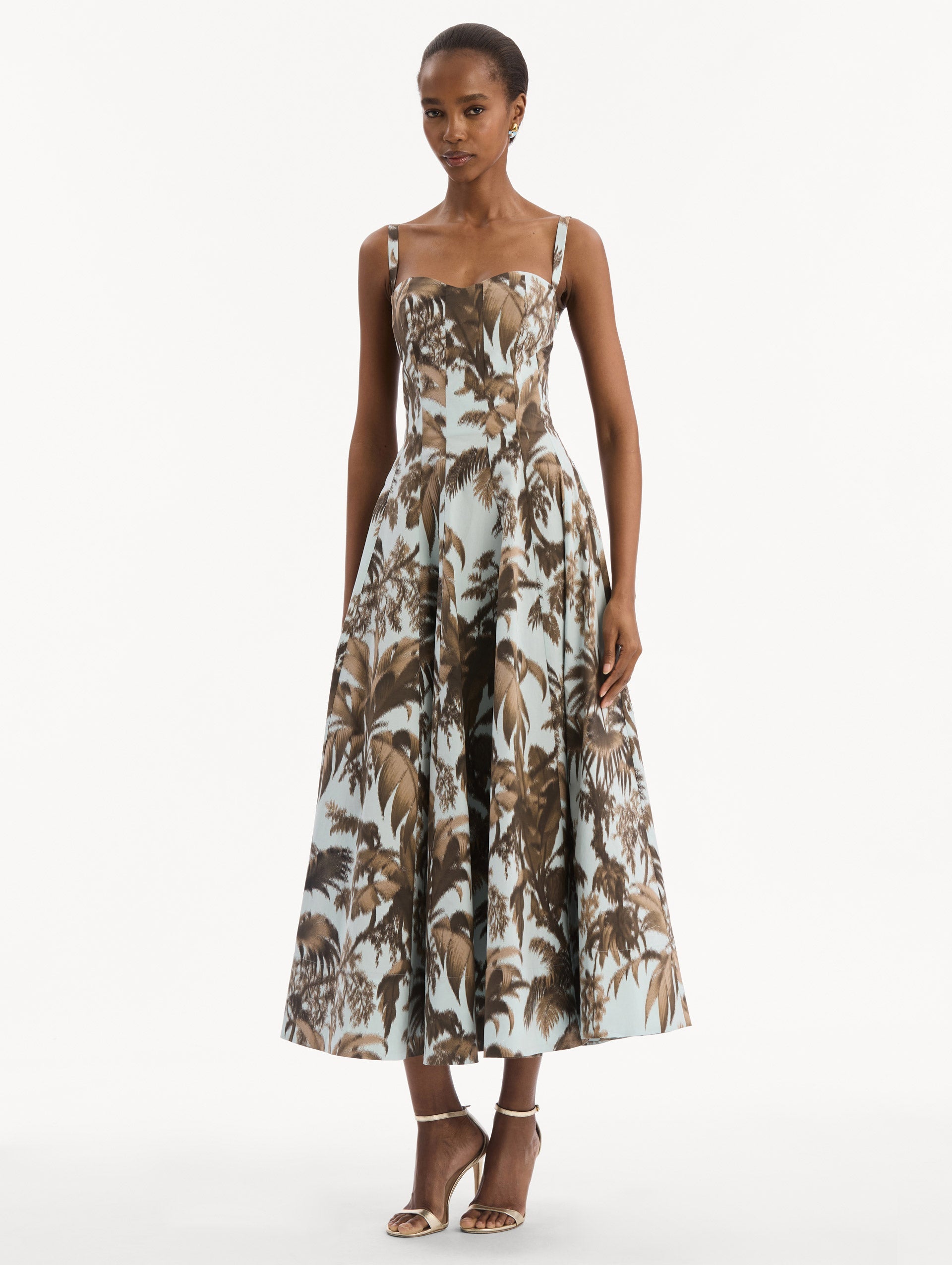 The model is wearing the Chiné Palm Leaves Cotton Poplin Dress in light blue printed with brown palm leaves. Featuring a subtle sweetheart neckline with tank straps, a softly structured bodice, and flowing midi skirt. Front View.