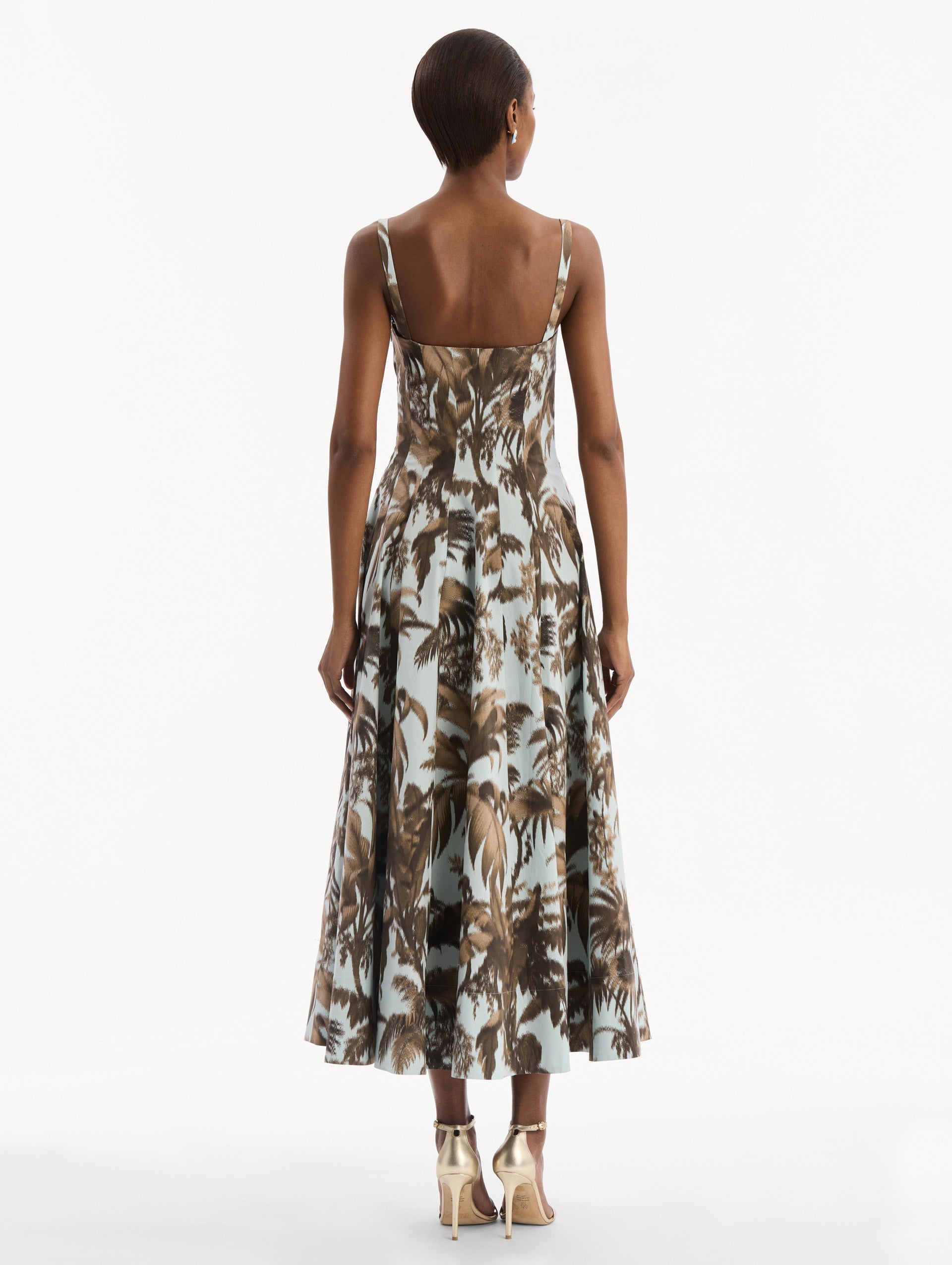 The model is wearing the Chiné Palm Leaves Cotton Poplin Dress in light blue printed with brown palm leaves. Featuring a subtle sweetheart neckline with tank straps, a softly structured bodice, and flowing midi skirt. Back View.