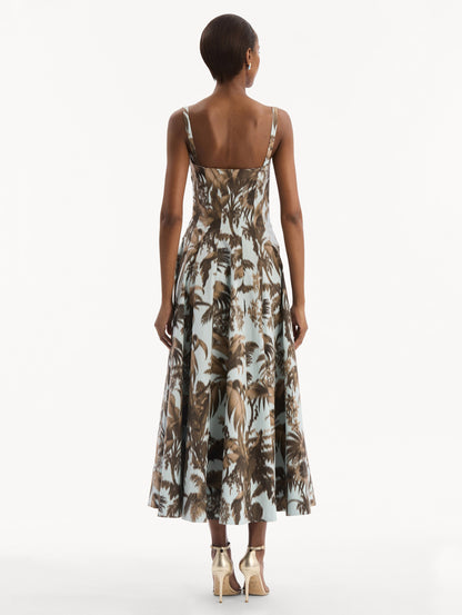 The model is wearing the Chiné Palm Leaves Cotton Poplin Dress in light blue printed with brown palm leaves. Featuring a subtle sweetheart neckline with tank straps, a softly structured bodice, and flowing midi skirt. Back View.