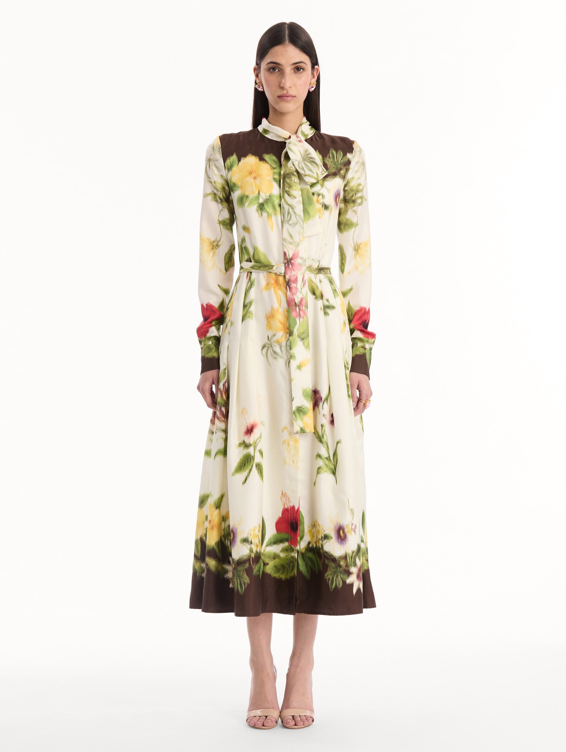 The model is wearing the Chiné Dominican Mixed-Floral Silk Twill Dress. Featuring a vibrant array of Dominican florals, tie neckline, belt at waist, long sleeves, and a midi length. Front View.