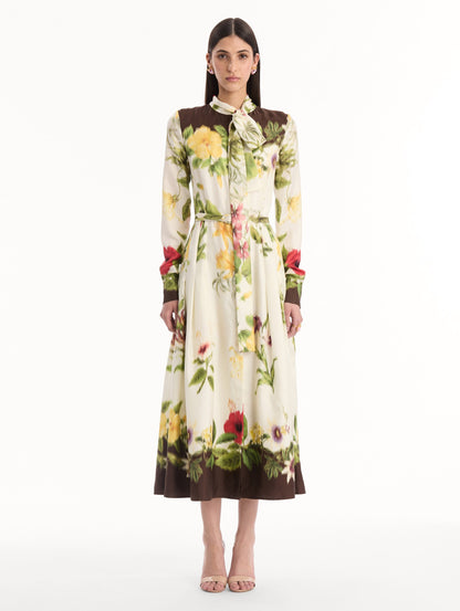 The model is wearing the Chiné Dominican Mixed-Floral Silk Twill Dress. Featuring a vibrant array of Dominican florals, tie neckline, belt at waist, long sleeves, and a midi length. Front View.