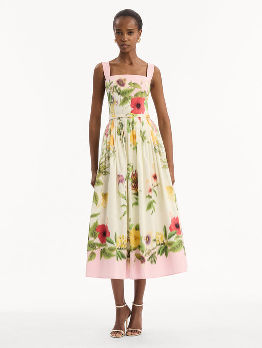 The model is wearing the Dominican Mixed-Floral Cotton Poplin Dress in ivory printed with an array of Dominican florals and light pink at the trims. Featuring a square neckline with tank straps, belt at waist, and pleated midi skirt. Front View.