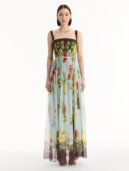 The model is wearing the Dominican Mixed-Floral Silk Chiffon Gown in light blue printed with an array of Dominican florals and brown at the trims. Featuring a pleated bodice, tank straps, tie at waist, and long, flowing skirt. Front View.