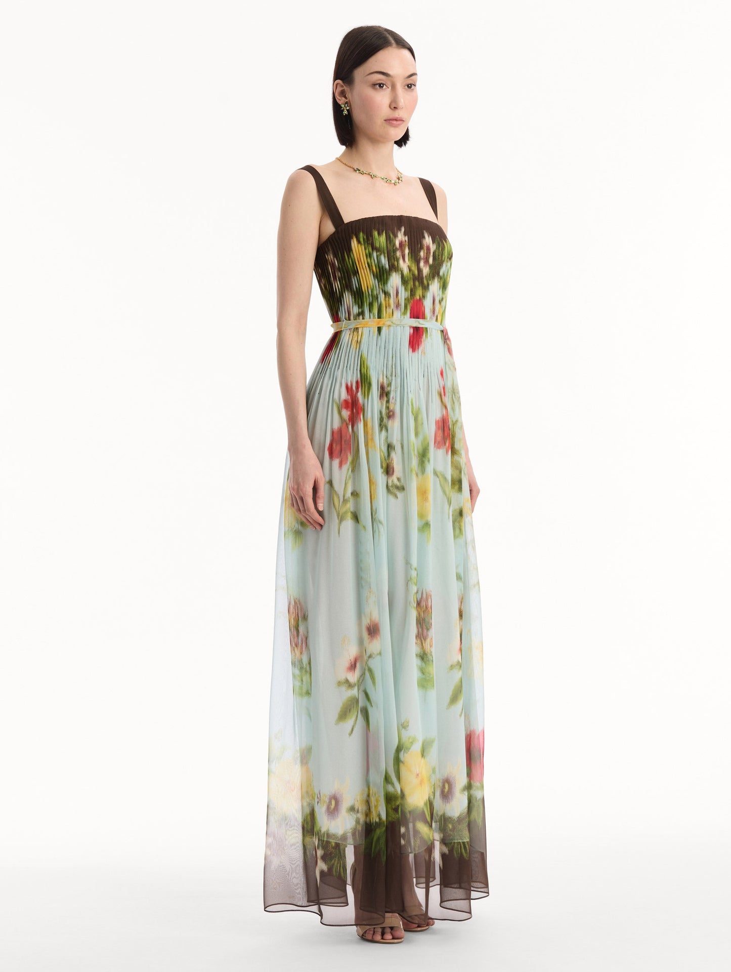 The model is wearing the Dominican Mixed-Floral Silk Chiffon Gown in light blue printed with an array of Dominican florals and brown at the trims. Featuring a pleated bodice, tank straps, tie at waist, and long, flowing skirt. Side View.