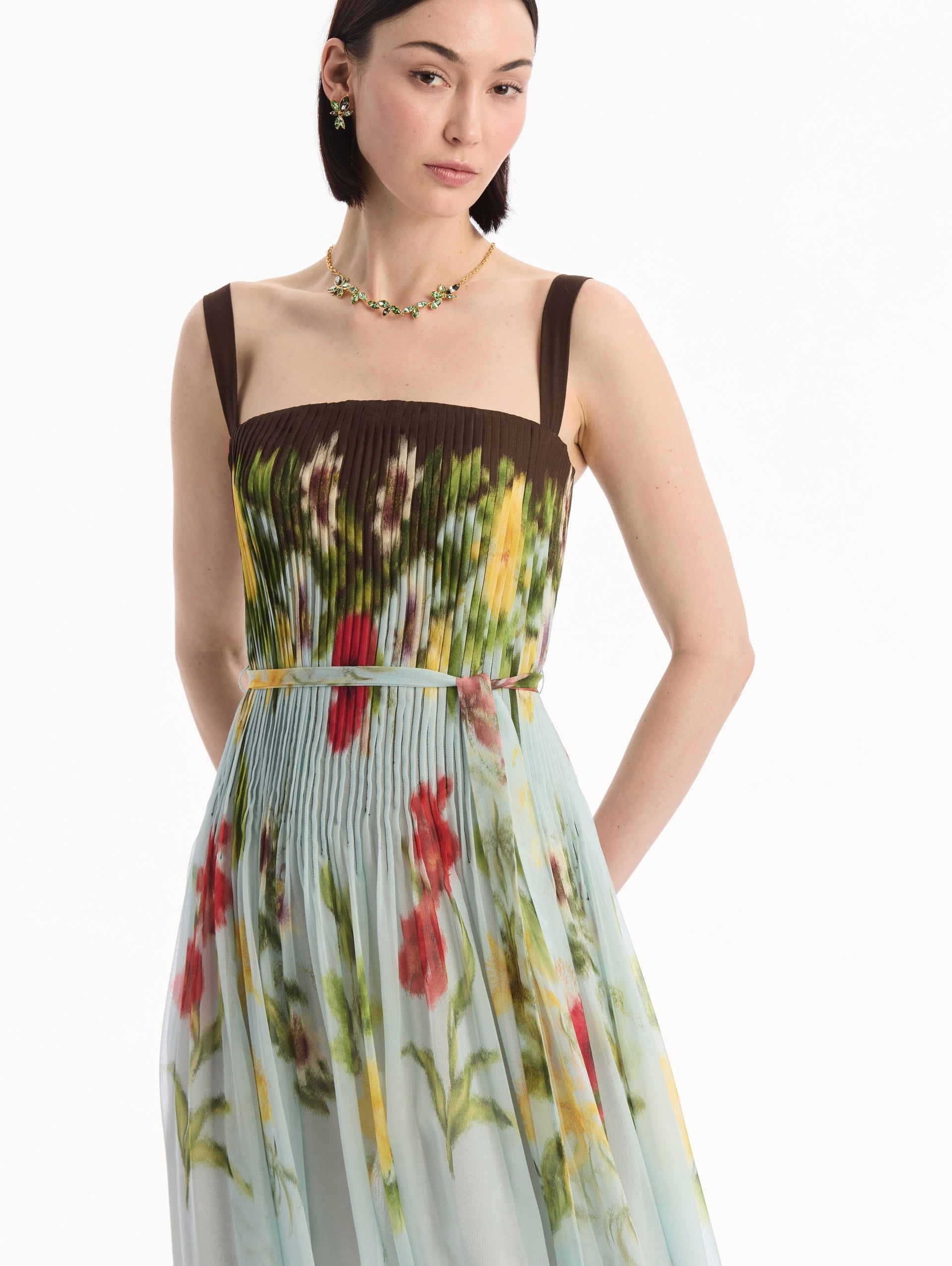 The model is wearing the Dominican Mixed-Floral Silk Chiffon Gown in light blue printed with an array of Dominican florals and brown at the trims. Featuring a pleated bodice, tank straps, tie at waist, and long, flowing skirt. Detail View.
