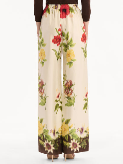 The model is wearing the Dominican Mixed-Floral Wide Leg Pant in ivory printed with an array of Dominican florals. Featuring wide legs and an elastic waistband. Back View.