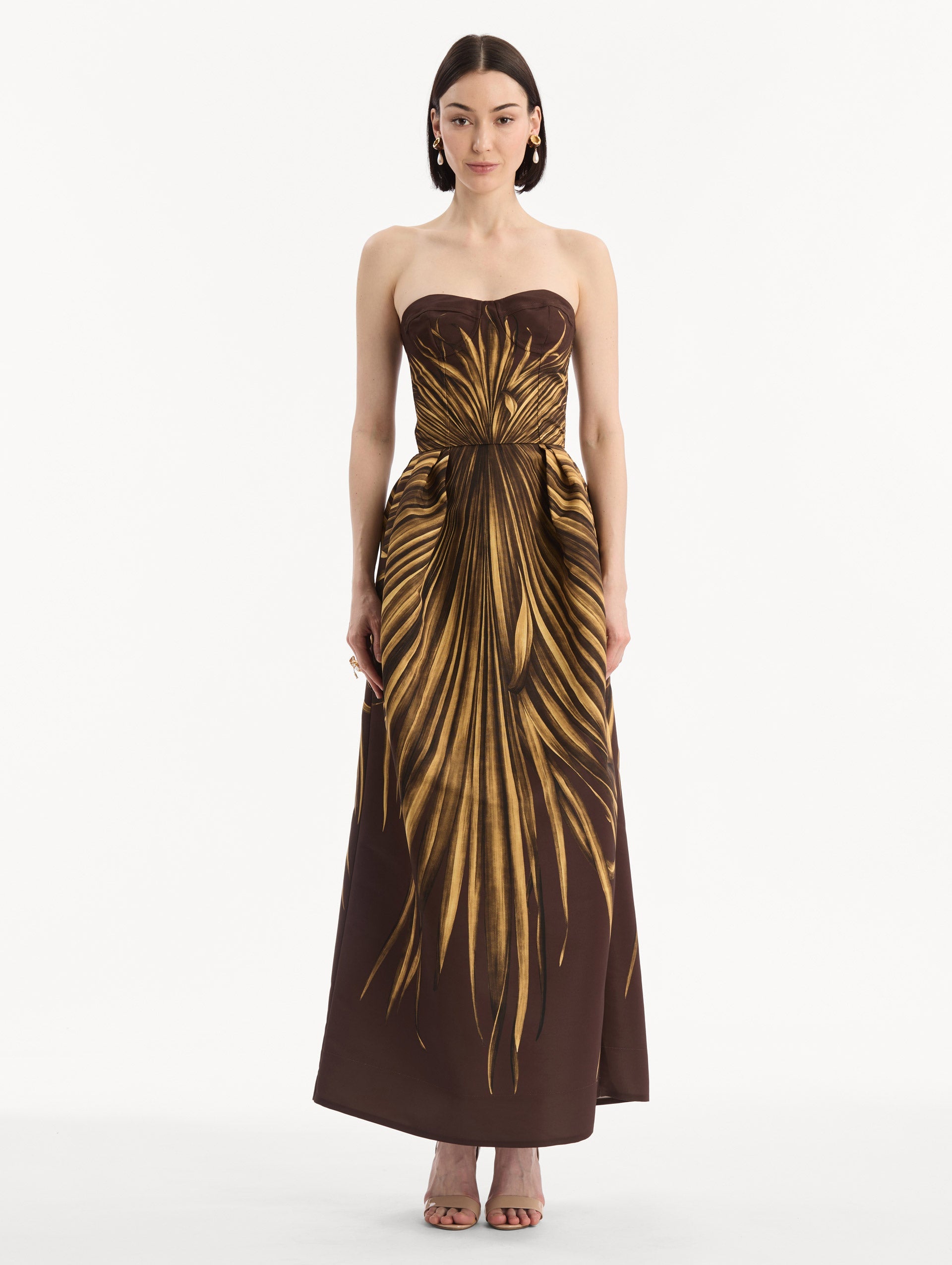 The model is wearing the Palm Leaves Faille Gown in brown printed with gold palm leaves. Featuring a strapless neckline, sculpted bodice, and long skirt. Front View.
