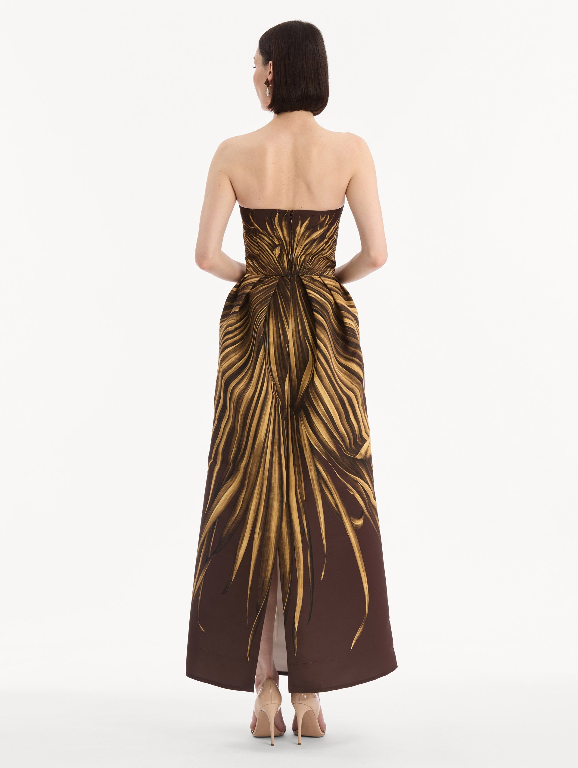 The model is wearing the Palm Leaves Faille Gown in brown printed with gold palm leaves. Featuring a strapless neckline, sculpted bodice, and long skirt. Back View.