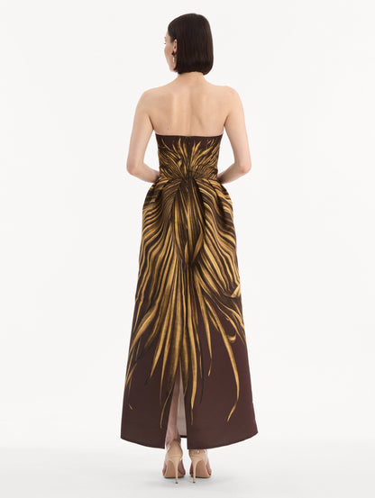The model is wearing the Palm Leaves Faille Gown in brown printed with gold palm leaves. Featuring a strapless neckline, sculpted bodice, and long skirt. Back View.