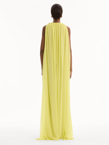 The model is wearing the Hibiscus-Detail Silk Chiffon Caftan in a light yellow-green color. Featuring a draping, gathered silhouette, long skirt, and sculptural hibiscus details at the shoulders. Back View.