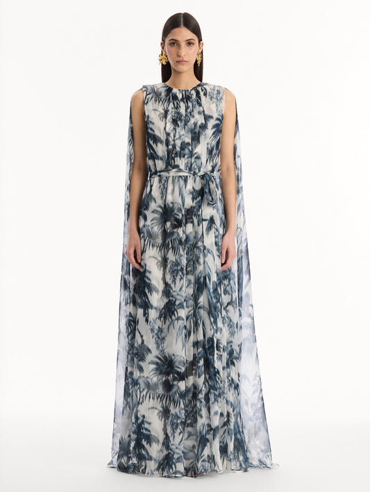 The model is wearing the Chiné Palm Leaves Silk Caftan White with Navy printed palm leaves. Featuring a gathered neckline, cape at back, flowing silhouette, and tie at waist. Front View.