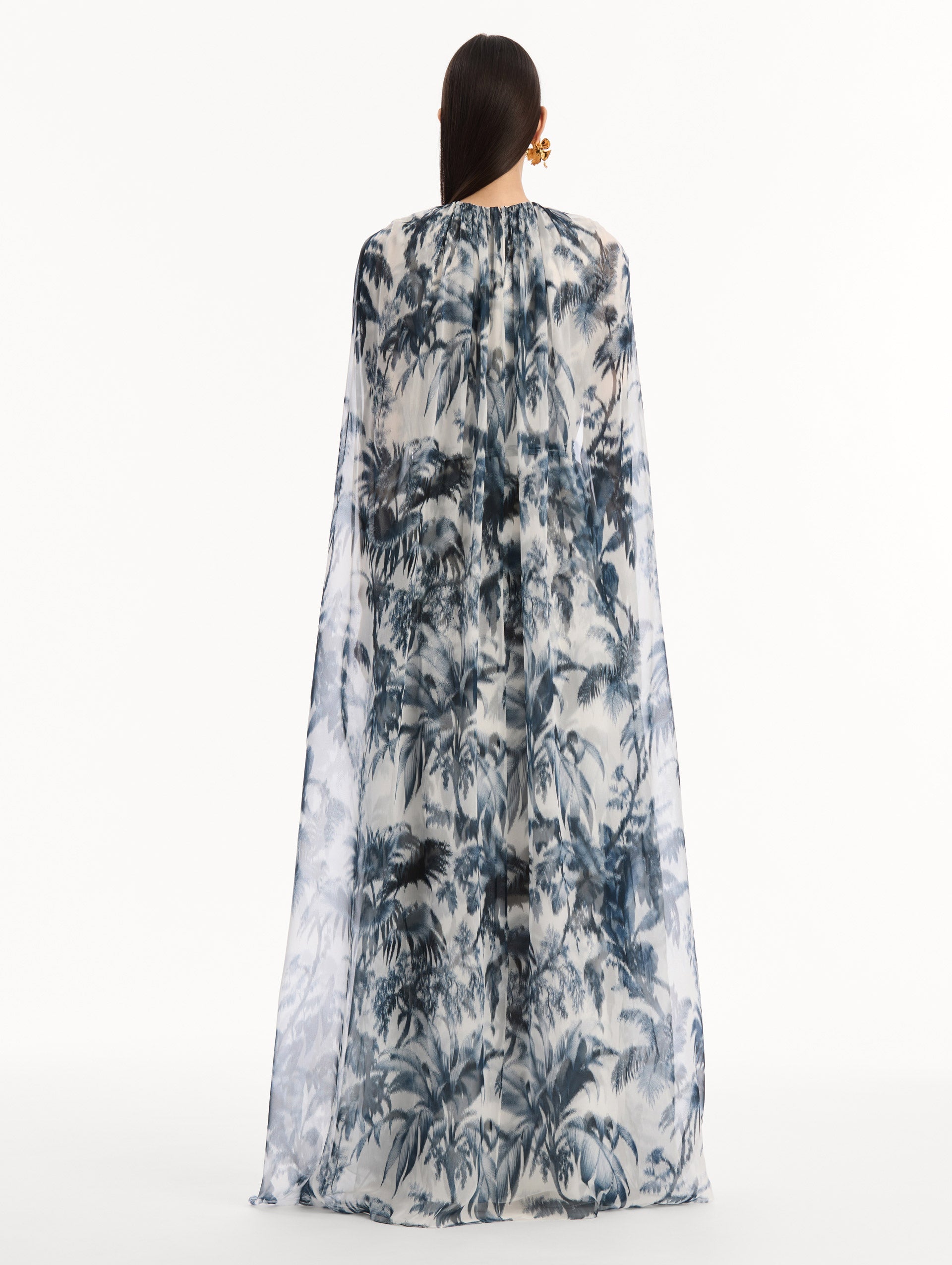 The model is wearing the Chiné Palm Leaves Silk Caftan White with Navy printed palm leaves. Featuring a gathered neckline, cape at back, flowing silhouette, and tie at waist. Back View.