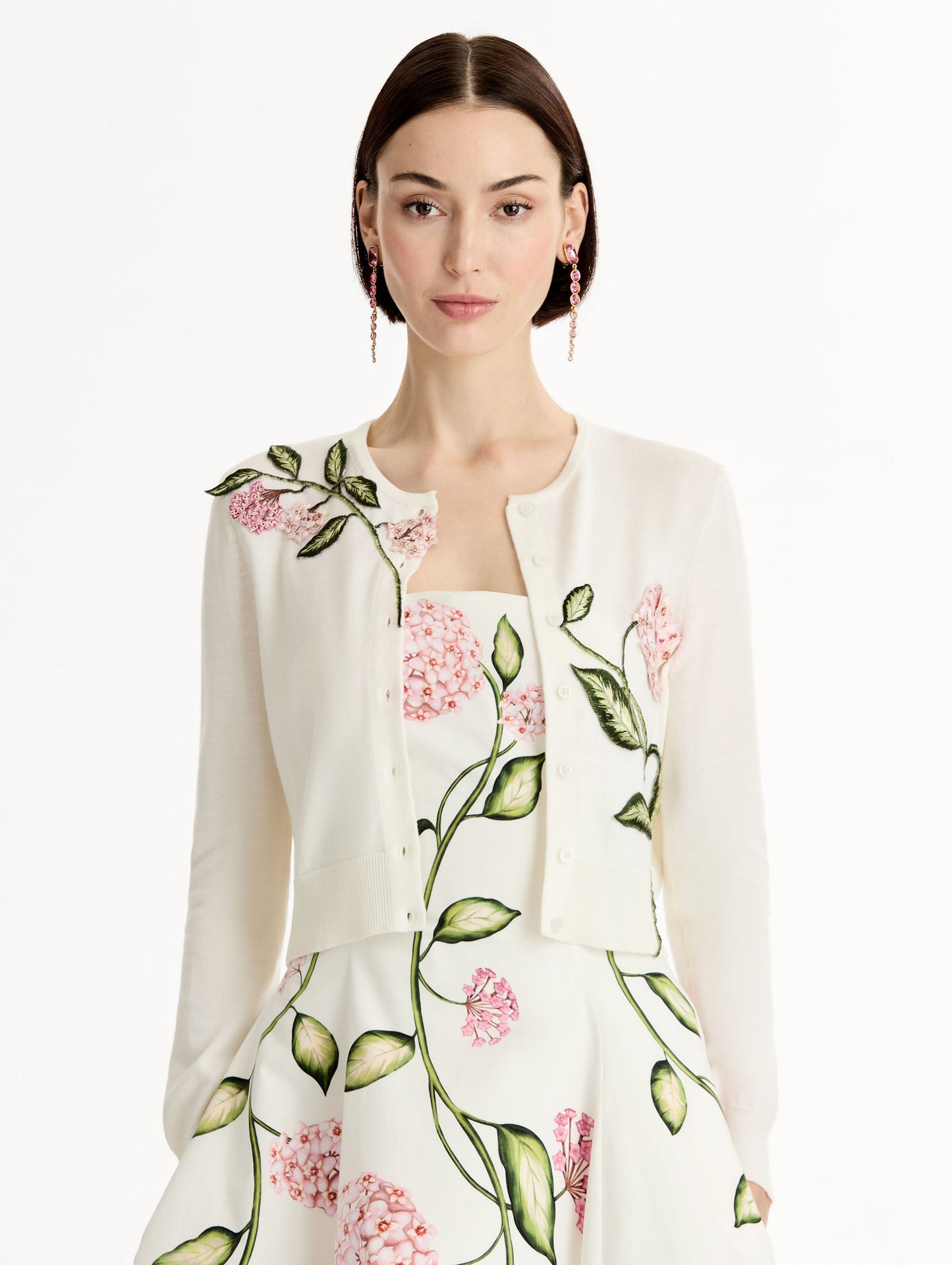 Ivory cropped cardigan with pink flower detailing stitched along the neckline and shoulder. Classic button closure. Crew Neck. Front View.