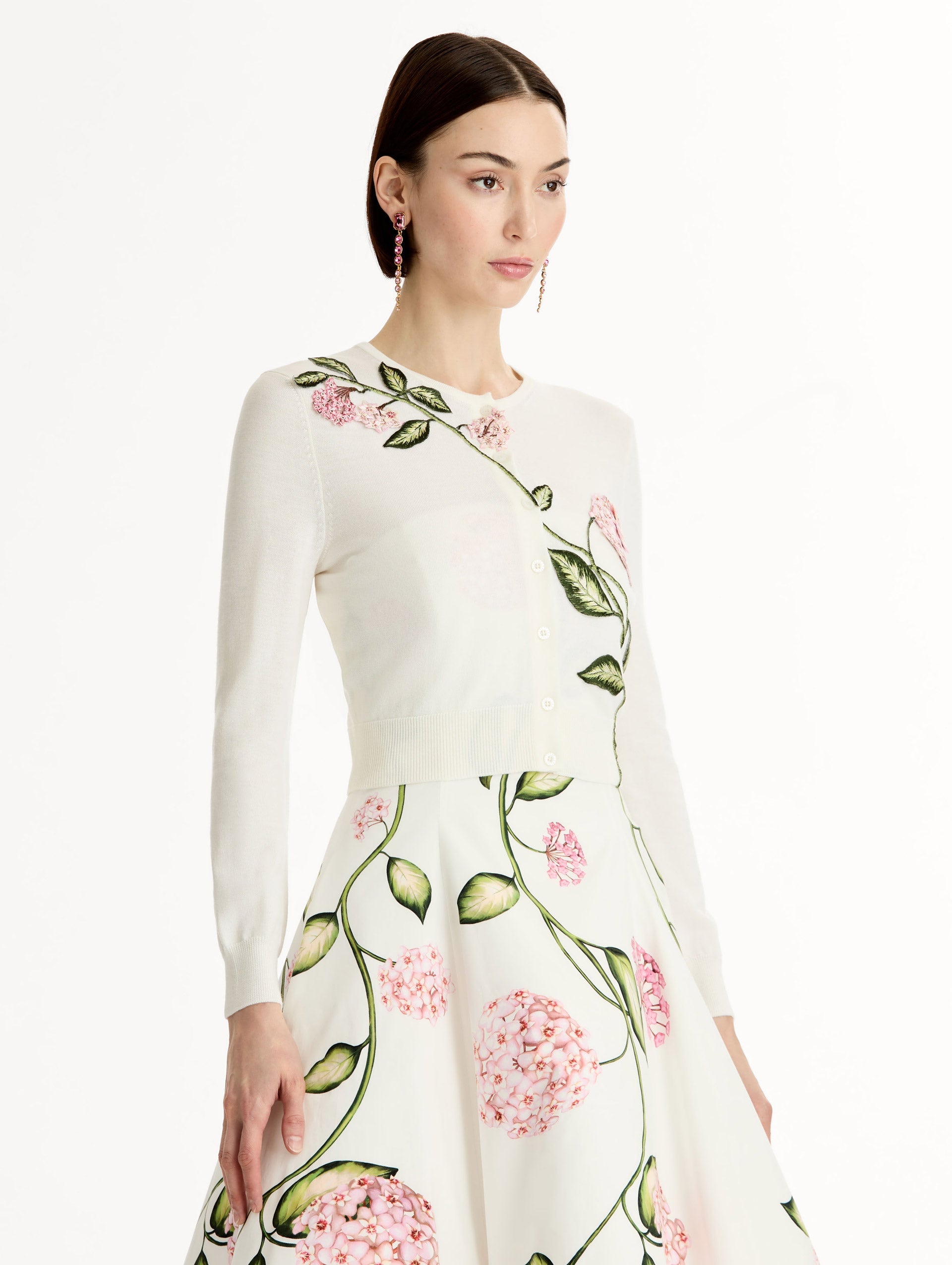 Ivory cropped cardigan with pink flower detailing stitched along the neckline and shoulder. Classic button closure. Crew Neck. Side View.