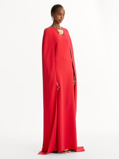 The model is wearing the Brooch Embroidered Cape-Back Caftan in the color red featuring draped sleeves and a floral brooch at center neckline. Side View.