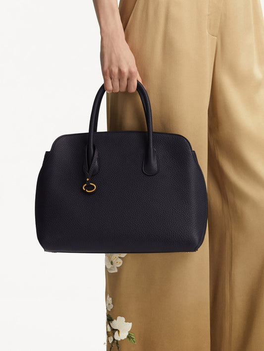 The model is holding the Ivy top handle handbag in black, paired with some khaki pants. The bag comes with the Oscar O logo as a tag around the handle.