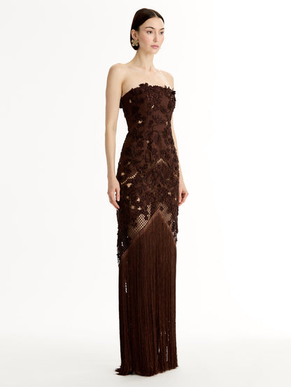 Mahogany strapless floor length gown with Porcelain flower threadwork in sculptural cutouts with glimpses of skin. Intricate lattice motifs. The silhouette elongates before cascading into a curtain of fringe. Side View.