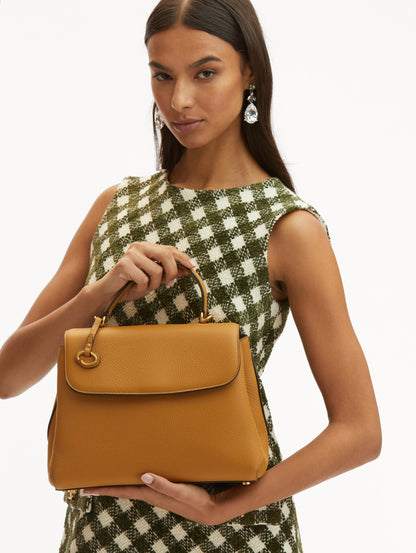 Model in plaid dress holding The Poppy bag in honey with O logo tag around the top handle. Front View