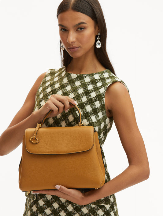 Model in plaid dress holding The Poppy bag in honey with O logo tag around the top handle. Front View