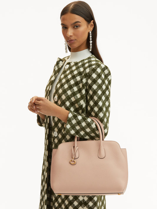 Model with plaid green and white coat holding The Ivy leather handbag in beige, with O logo tag around the handle Front View