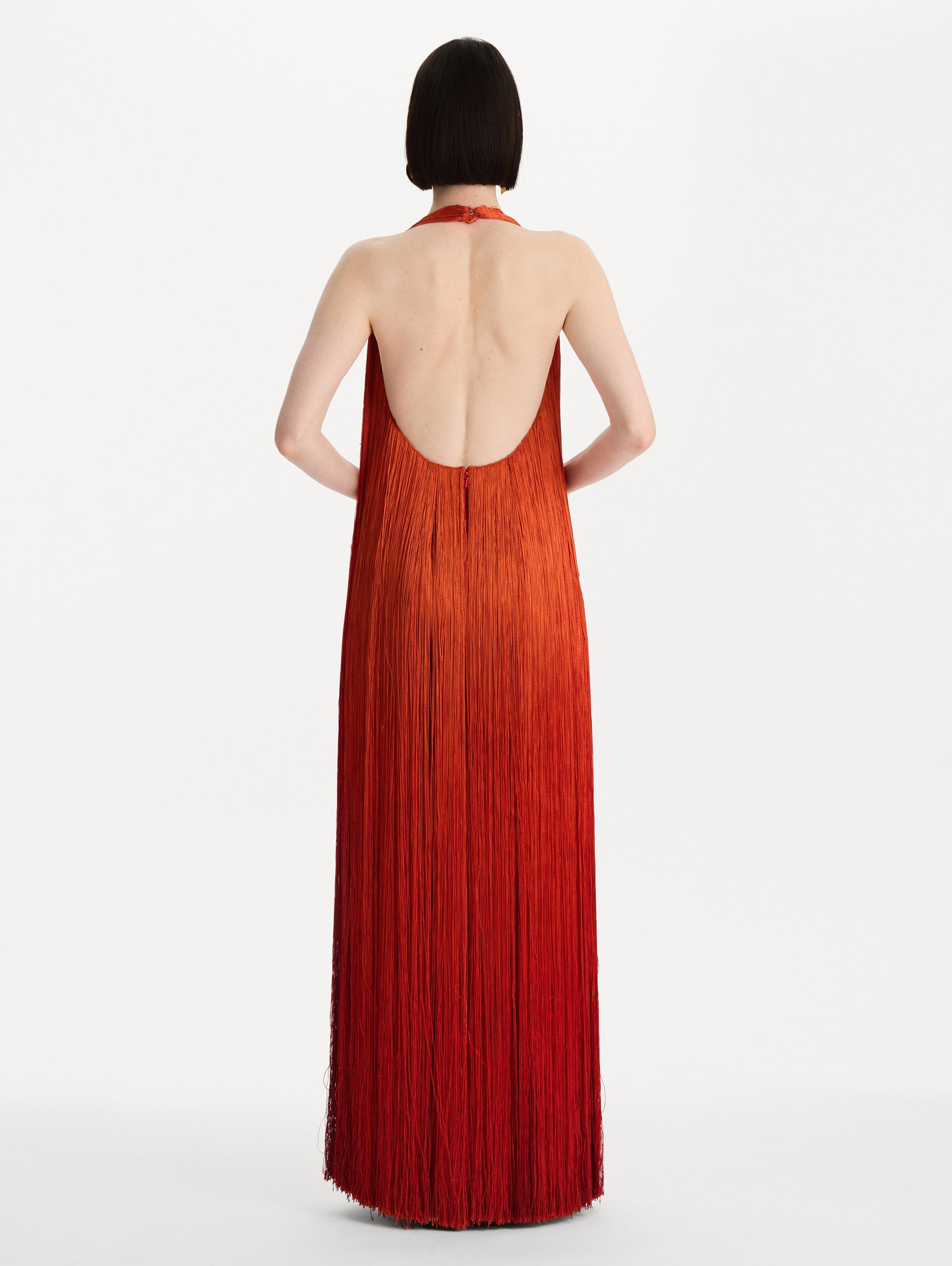 The model is wearing the Ombré Fringe Gown in Paprika Red. Featuring a deep, halter neckline, and full fringe silhouette. Back View.