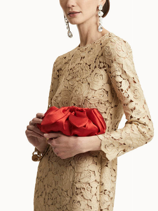 Model in beige long sleeve guipure dress and crystal drop earrings, holds a red satin clutch with a bow in the top.